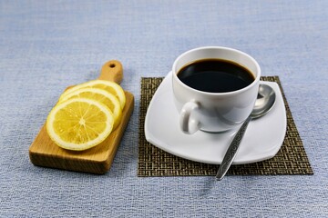 White cup of black coffee on a napkin mat and lemon slices on a small cutting board.