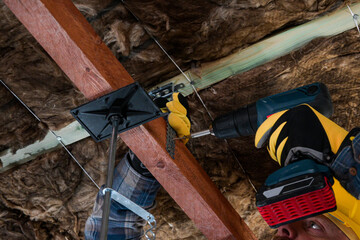 worker in overalls and helmet with yellow gloves drives a screw into the ceiling with a cordless drill - close up view
