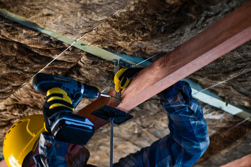 worker in overalls and helmet with yellow gloves drives a screw into the ceiling with a cordless...