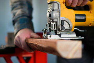 worker in overalls and wearing yellow gloves sawing wood with a reciprocating electric wood saw - close-up view and blurred background