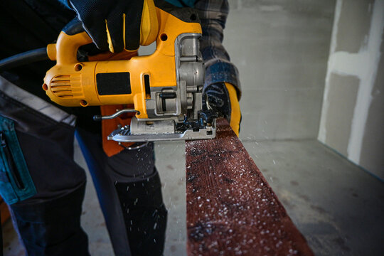 worker in overalls and wearing yellow gloves sawing wood with a reciprocating electric wood saw - close-up view and blurred background