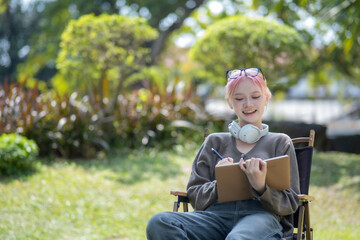 Fototapeta premium Young Happy Asian artist woman drawing book and wearing headphone, the artist paints in the garden
