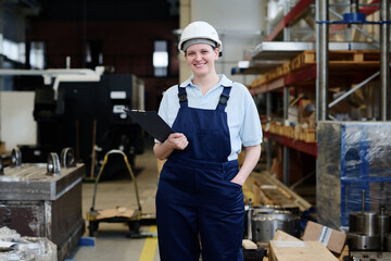 Fototapeta na wymiar Medium long shot of cheerful young Caucasian female factory worker holding clipboard smiling at camera