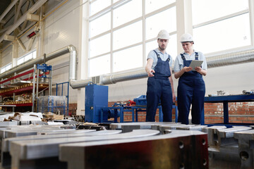 Wide shot of Caucasian man wearing hardhat demonstrating metal object produced in factory to his...