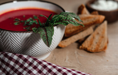 beetroot tomatoes soup in white bowl with handles served with crunchy triangle toasted bread, green beet leaves, green garlic, rustic cloth and wooden background