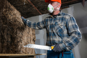 a worker in overalls, gloves and a respirator holds a cut piece of glass wool in his hand, a knife in one hand