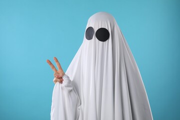 Funny ghost. Woman covered with white sheet showing V-sign on light blue background