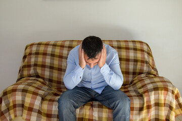 Image of a man sitting on a sofa while covering his ears and not wanting to hear anyone. Anxiety and depression problems.
