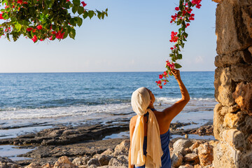 Beautiful woman with headscarf and blue sarong picking up a branch of bougainvillea to smell its...