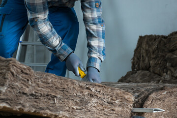 a worker in overalls, gloves and a respirator cuts glass wool with a large knife