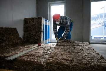 a worker in overalls, gloves and a respirator cuts glass wool with a large knife
