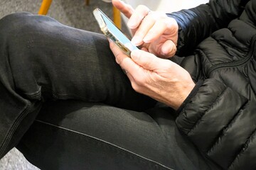 Auf Stuhl sitzender Mann mit schwarzer Jeanshose und schwarzer Steppjacke hält Handy in Händen in...