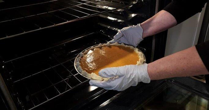Gloved hands carry an unbaked pumpkin pie from kitchen counter to a pre-heated oven for baking.