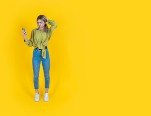 Irritated confused young woman, full body length front view of irritated confused young woman. Hold use smartphone on hand looking screen scratching her head. Reading bad news. Yellow background.