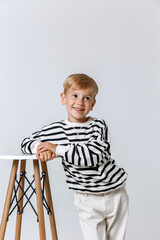 Little smiling kid boy in striped sweatshirt and white trousers posing at studio as a fashion model.