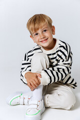 Little smiling blonde kid boy in striped sweatshirt and white trousers sitting and posing at studio