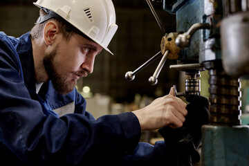 Side view medium closeup of young man wearing hardhat repairing industrial equipment in workshop