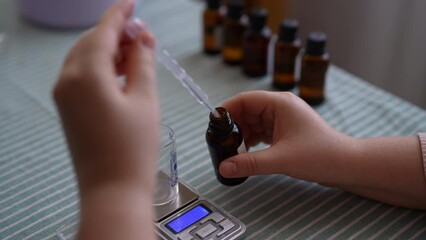 Closeup rear view of unrecognizable woman dropping essential oil into glass bottle using pipette sitting at table. Process of making handmade natural scented candle at workshop. Concept of handicraft