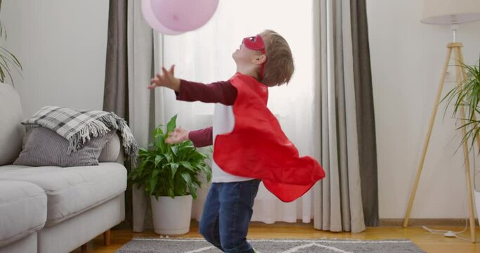 Child in superhero costume with red cape and mask playing with balloons in home
