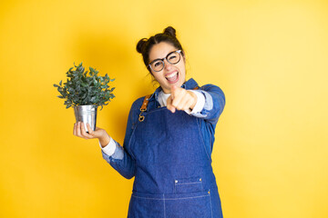 Young caucasian gardener woman holding a plant isolated on yellow background laughing at you,...