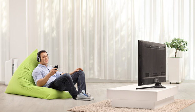 Young Man Sitting On A Beanbag Armchair And Listening To Music With Headphones And Smartphone In Front Of Tv