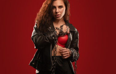 Sexy red-haired curly girl with beautiful makeup in a black leather jacket on a red background. She's got chains on her hands