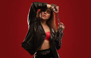 Sexy red-haired curly girl with beautiful makeup in a black leather jacket on a red background. She's got chains on her hands