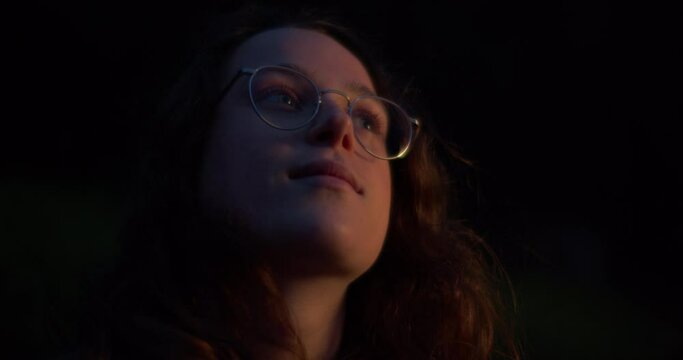 Cinematic shot of young woman looking at the night sky and smiling. Light reflects in the girls glasses. Romantic and dreamy mood. Enjoying freedom and youth. Pensive and introvert girl alone. 