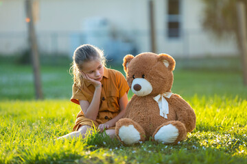 Sad lonely child girl spending time together with her teddy bear friend outdoors on sunny backyard....