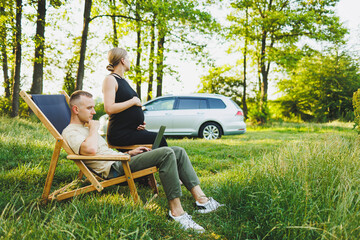 Fototapeta premium A family couple, a man and a woman, are relaxing in nature and working remotely with a laptop. Picnic of loving couples with work online. A pregnant woman rests in nature with her husband