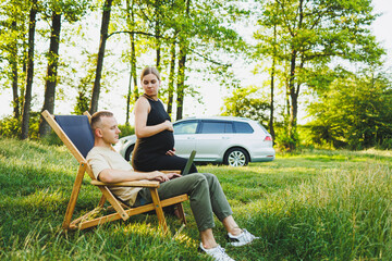 Fototapeta premium A happy couple relaxes in folding chairs near the car, a man uses a laptop for work or checking social networks, his wife treats him to a croissant. Pregnant woman with man in nature.