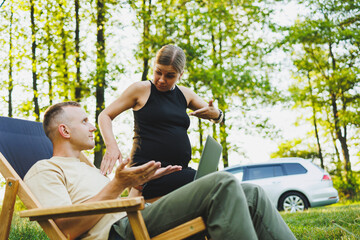 A happy couple relaxes in folding chairs near the car, a man uses a laptop for work or checking social networks, his wife treats him to a croissant. Pregnant woman with man in nature.