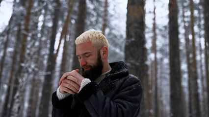 Stylish hipster man in pine tree winter forest rubbing his cold red face with his hands. Media. Portrait of a man outdoors spending time alone in winter woods.