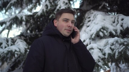 A young man speaks on the phone while standing against a background of spruce in winter. A man with a phone against a background of snowy spruce branches.