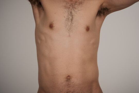 The torso of a shirtless man with his arm raised, showing muscle definition on his slim physique. He has olive skin and a small amount of natural armpit hair, chest hair, and belly button hair.