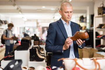 Portrait of focused man choosing formal shoes in store, looking with interest at stylish brown...