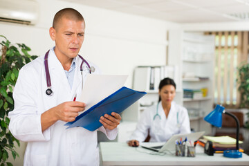 Portrait of experienced doctor standing in medical office with papers in hands, focused on studying of clinical diagnosis of patient