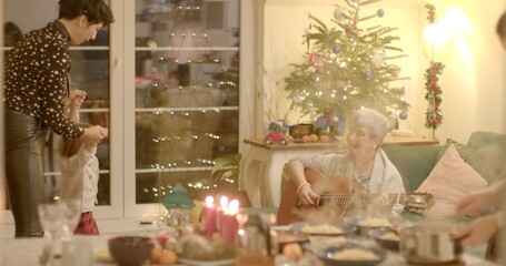 Cozy Christmas celebration at home unfolds as mother and daughter joyfully dance to the musical accompaniment provided by their grandmother guest. Festive atmosphere filled with love and togetherness.