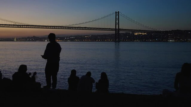 Silhouette of man feeding woman and people taking photos of sunset in Jardim do Rio park waterfront in Cacilhas, Alamda in Lisbon, Portugal