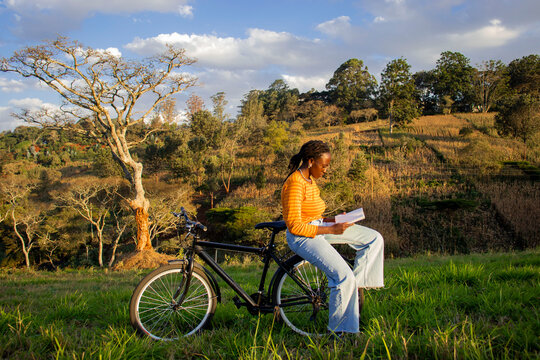 Young Black woman reading a book on a bicycle in a field
