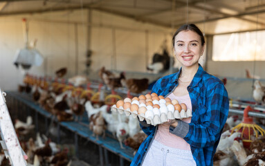 Happy successful female owner of poultry farm engaged in breeding of laying hens standing in chicken coop, holding carton tray of fresh organic eggs © JackF