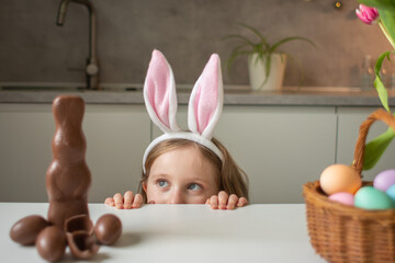 Cute little girl wearing bunny ears eating chocolate Easter rabbit. Kid playing egg hunt on Easter. Adorable child celebrate Easter at home