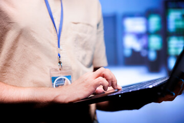 Worker in front of blurry background supercomputers delivering web content to online customers. Repairman in server room using laptop to make sure critical systems are working errorless, close up