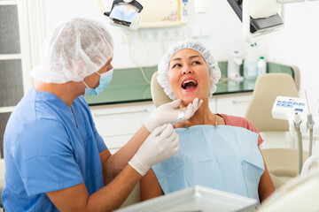 Qualified man dentist in a protective mask conducts an examines of a woman patient sitting in a...