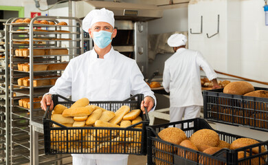 Baker in face mask for viral protection holding crate with bread, industrial kitchen of bakery on background