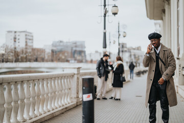Elegant man in a stylish overcoat and beret talking on his smart phone on a city street, with pedestrians in the soft-focus background.