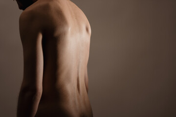 A nude man stands alone in a dimly lit studio, slouching and looking down. He has his back turned...