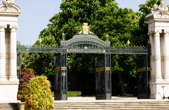 Madrid, Spain. 7 May 2022. Black metal gate with white columns at entrance to Retiro Park. Botanical Garden on a sunny spring day when the trees are in bloom. People walking outdoors, riding bicycles.