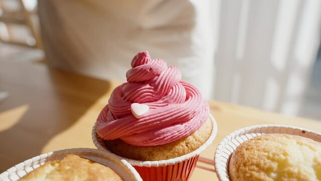 A woman decorates cupcakes, placing heart-shaped sugar decorations on top of pink frosting hats.