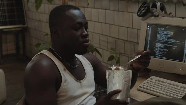 Young Black programmer eating meal from foil pouch and typing code on old computer while surviving in post apocalypse shelter